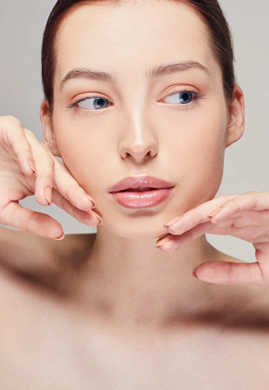 Sensual girl with brown hair clean fresh skin naked shoulders touches her face by her hands posing at gray studio background, looking straight, a model with light nude make-up, copy space