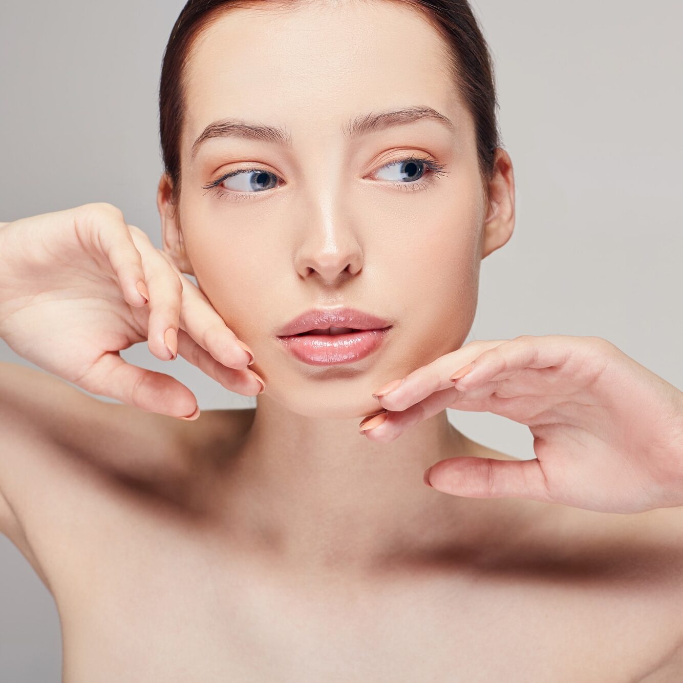 Sensual girl with brown hair clean fresh skin naked shoulders touches her face by her hands posing at gray studio background, looking straight, a model with light nude make-up, copy space