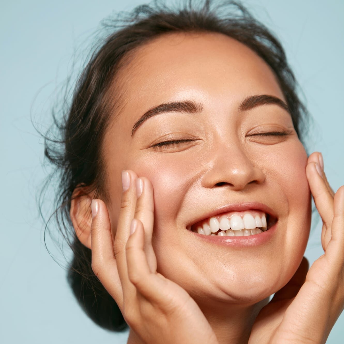 Skin care. Woman with beauty face touching healthy facial skin portrait. Beautiful smiling asian girl model with natural makeup touching glowing hydrated skin on blue background closeup