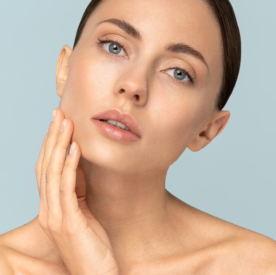 Studio portrait of young woman with natural makeup, combed hair, touching well-groomed pure skin on face, looking at camera, blue background, banner. Beauty, facelift, anti-aging cosmetic procedure.